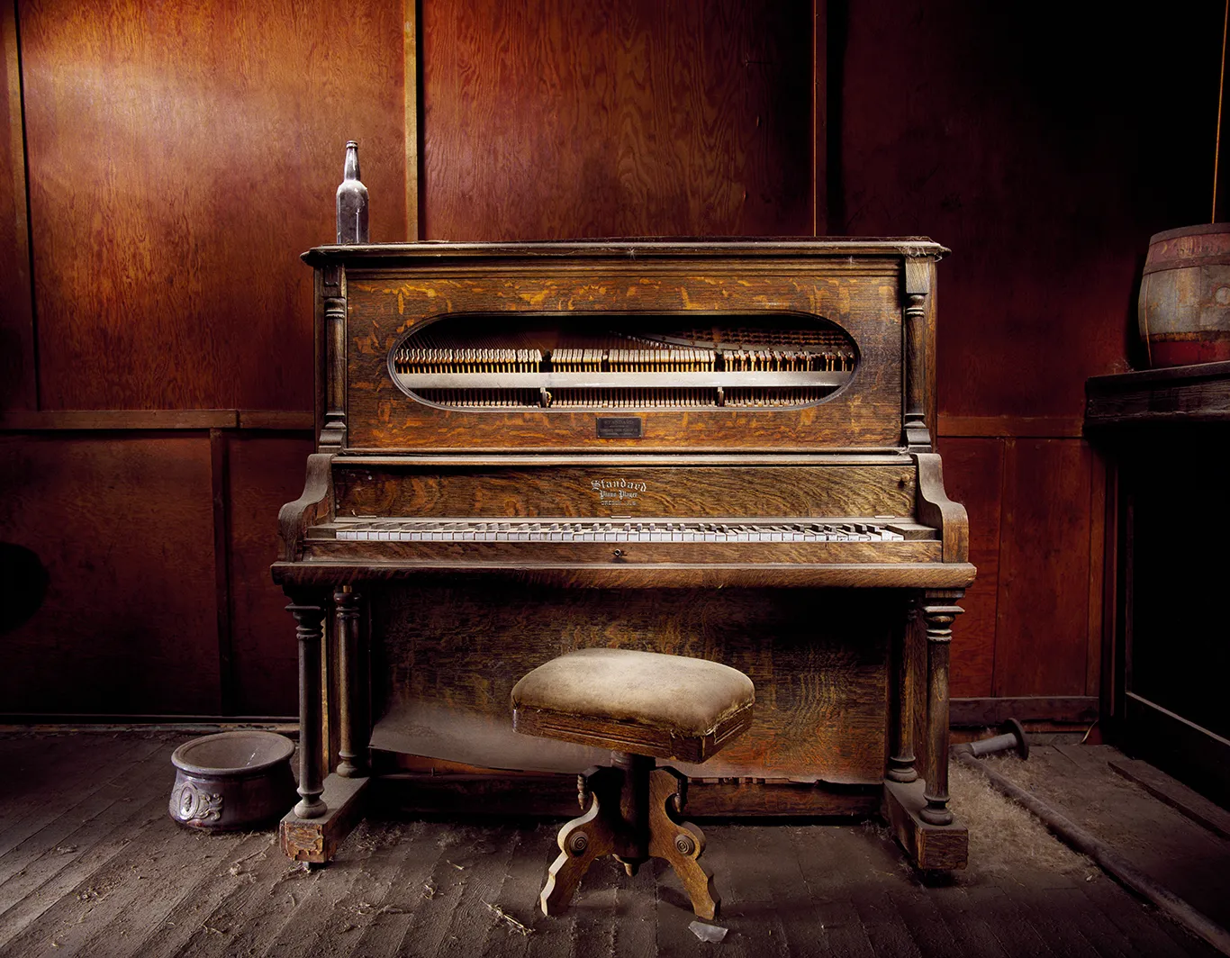 A dust-covered, vintage upright piano with weathered wood and an empty bottle resting on top of it, inside the abandoned Sam Leon Bar in the ghost town of Bodie