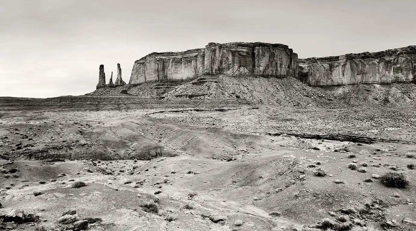 Schwarzweiß-Fotografie der Felsformation „Three Sisters“ im Monument Valley, drei schlanke Sandsteinnadeln vor weitem Horizont.