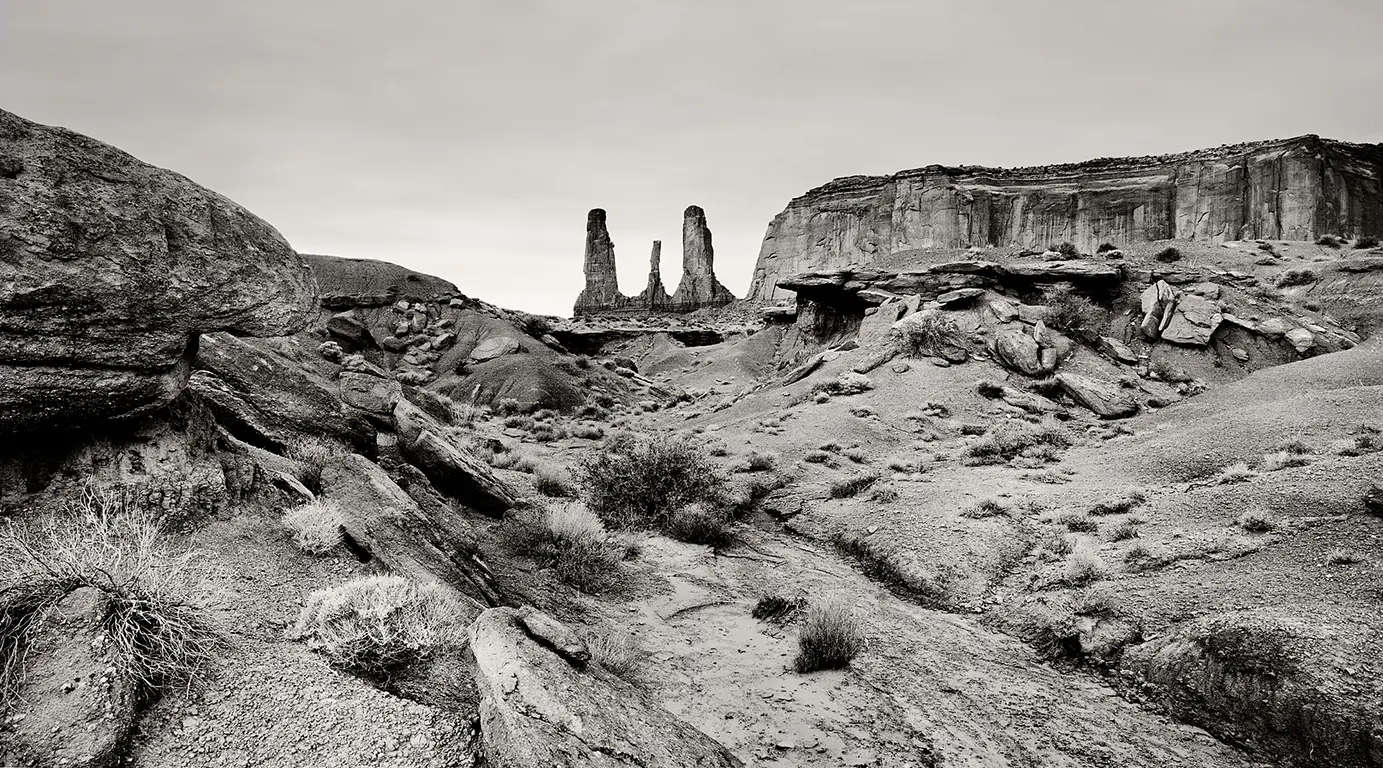 black-and-white photograph of the ‘Three Sisters’ rock formation in Monument Valley, showing three sandstone spires against the horizon, with a striking foreground of stones and scree